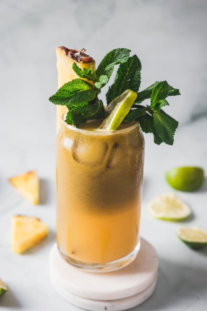 A tall glass filled with an iced brown and orange drink, garnished with a lime wedge, a pineapple slice, and fresh mint leaves. Slices of lime and pineapple are scattered on a light surface in the background.