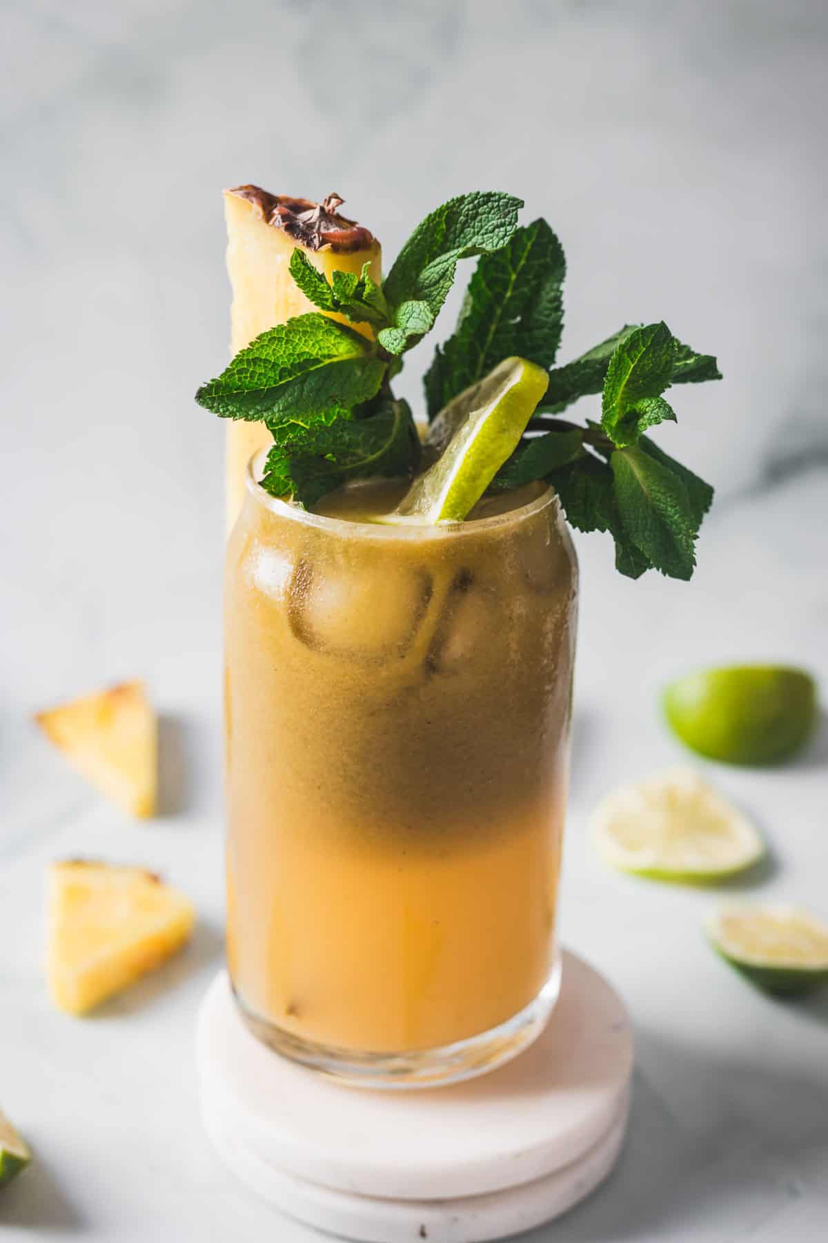 A tall glass filled with an iced brown and orange drink, garnished with a lime wedge, a pineapple slice, and fresh mint leaves. Slices of lime and pineapple are scattered on a light surface in the background.