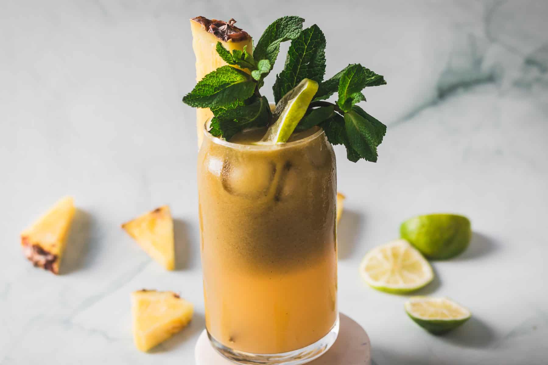 A tall glass filled with a yellowish Colombian Lulo Juice cocktail on ice, garnished with a pineapple wedge, lime slice, and fresh mint. Slices of pineapple and lime are scattered on a white marble surface in the background.
