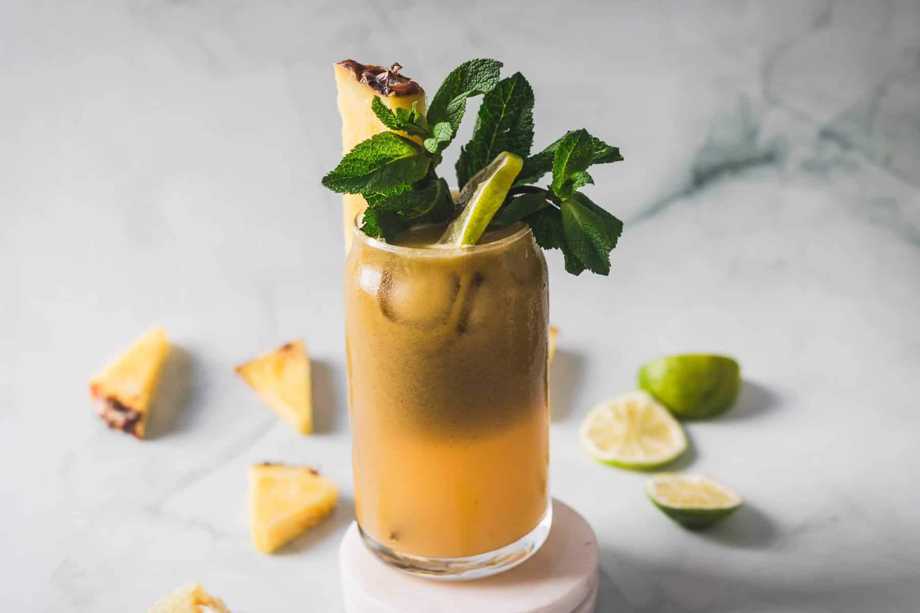 A tall glass of iced tropical drink garnished with a pineapple wedge, mint leaves, and a lime slice, sits on a round white stand. Sliced lime and pineapple pieces are scattered on a light marble surface.