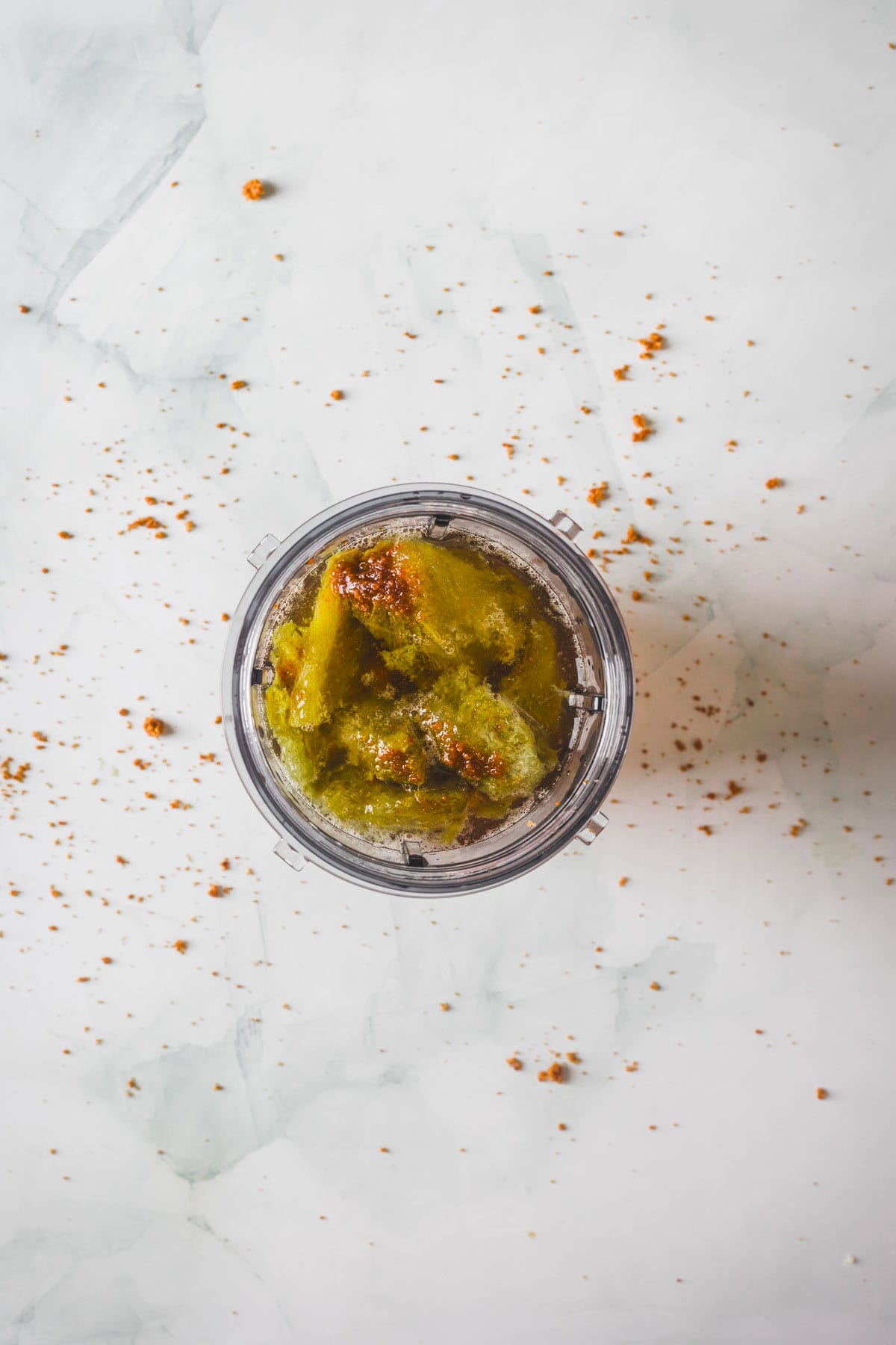A top-down view of a blender cup filled with chunky green ingredients and spices, ready to make Colombian Lulo Juice, placed on a white surface with brown spice scattered around.