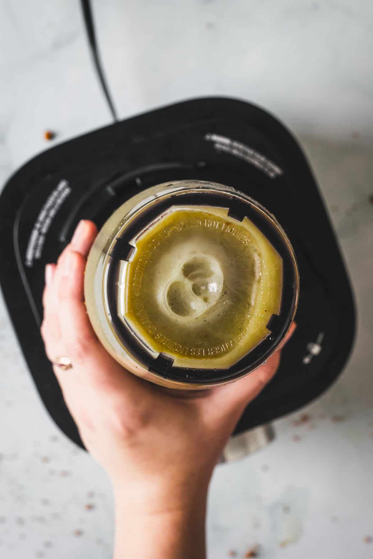A hand holds the lid of a blender filled with vibrant Colombian Lulo Juice, seen from above. The greenish smoothie swirls inside as the blender sits on a countertop, its base partially visible.