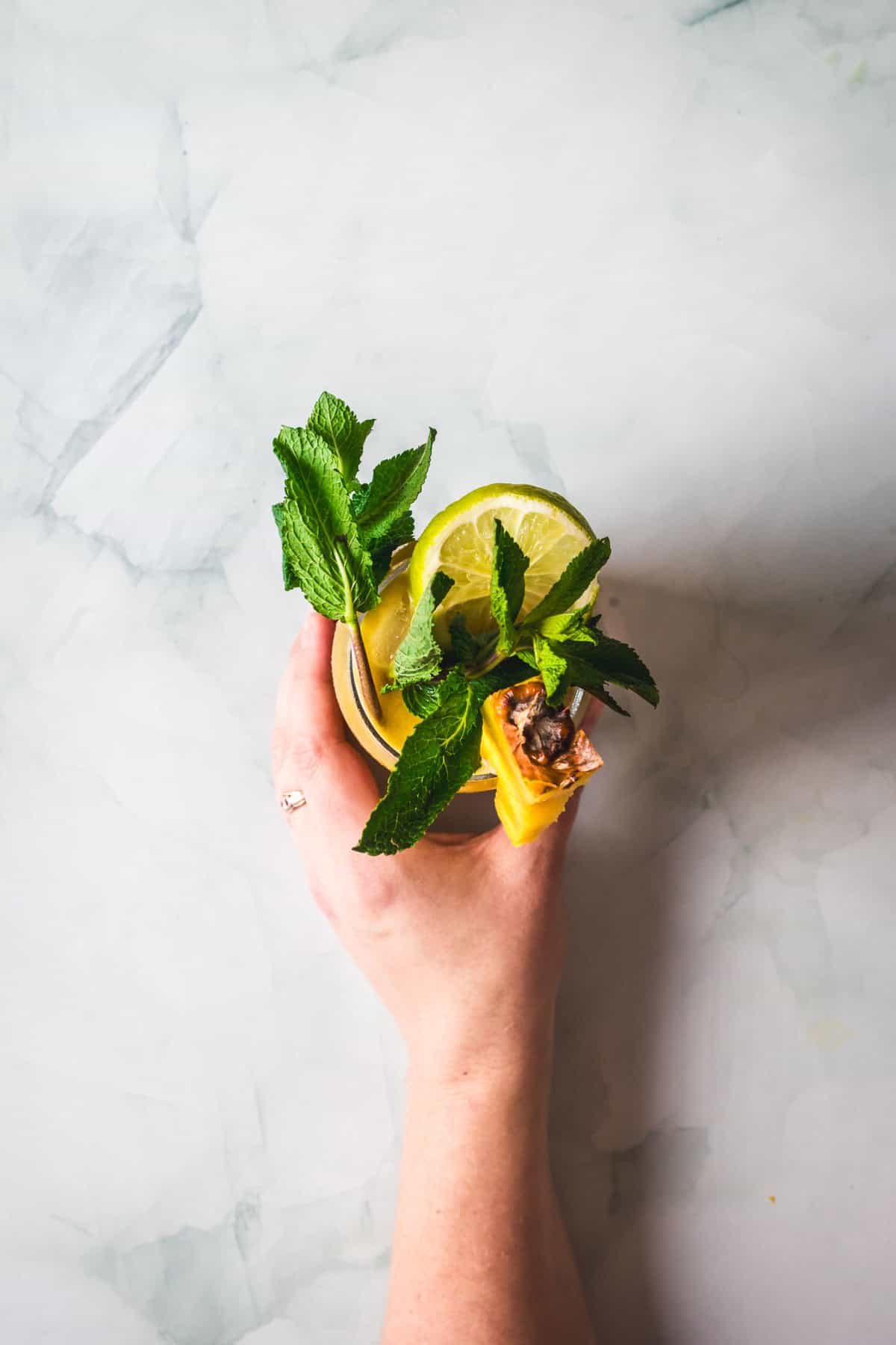 A hand holding a glass of Colombian Lulo Juice, garnished with fresh mint, lemon slices, and a small piece of honeycomb against a light, textured background.