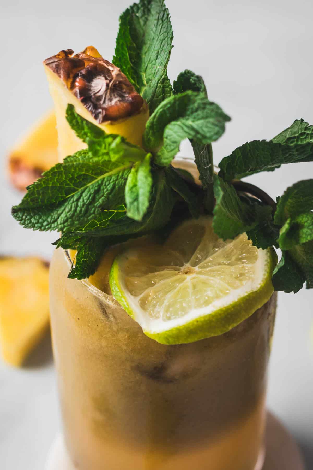 A close-up of a cold drink garnished with a slice of lime, a sprig of fresh mint, and a piece of grilled pineapple on the rim of the glass. The drink contains ice and is presented against a neutral background.