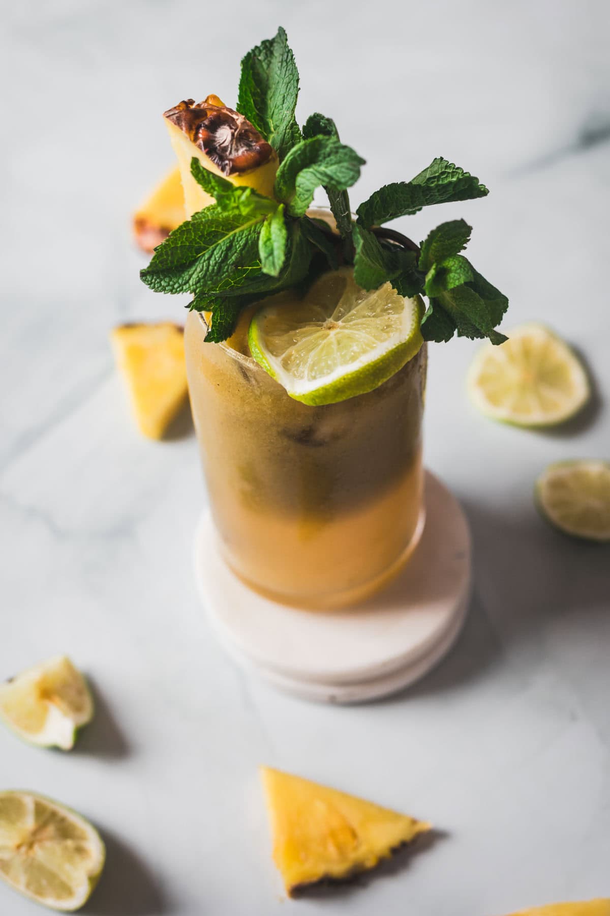 A tall glass filled with Colombian Lulo Juice, a vibrant yellow-orange drink, garnished with a lime slice, pineapple wedge, and fresh mint leaves. Extra lime slices and pineapple pieces are scattered on the light surface around the glass.