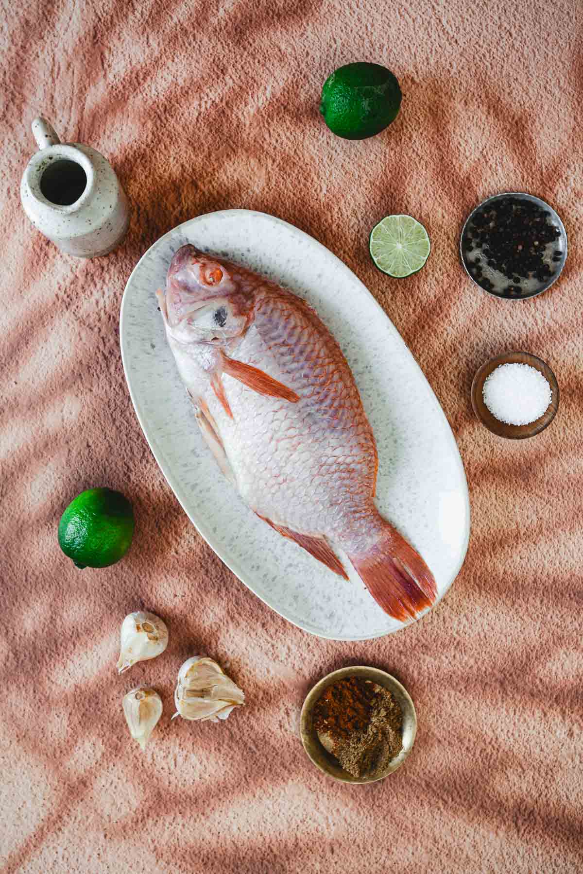 A whole raw fish for Colombian Whole Fried Mojarra Frita rests on an oval plate, surrounded by limes, a small pitcher, a bowl of spice mix, garlic cloves, pepper, and salt—all atop a textured pink surface.
