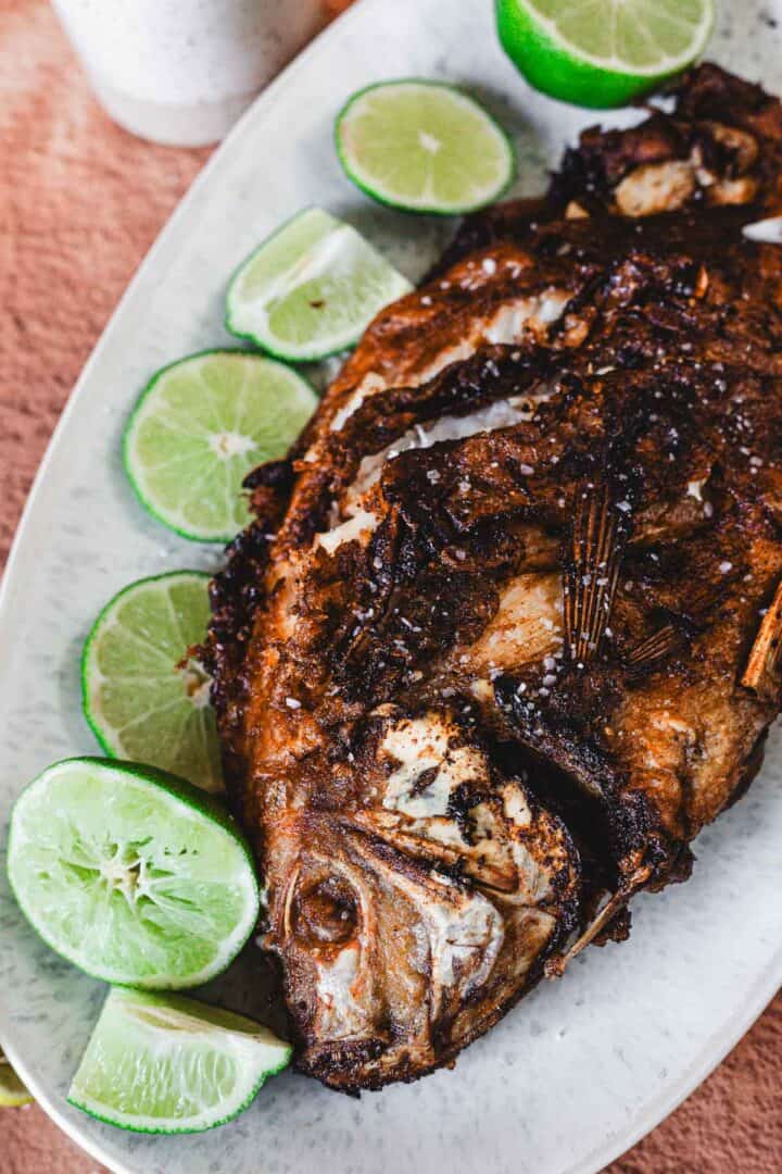 A Whole Fried Mojarra with crispy brown skin is served on an oval plate, garnished with several lime wedges—a classic Colombian favorite, also known as Mojarra Frita.