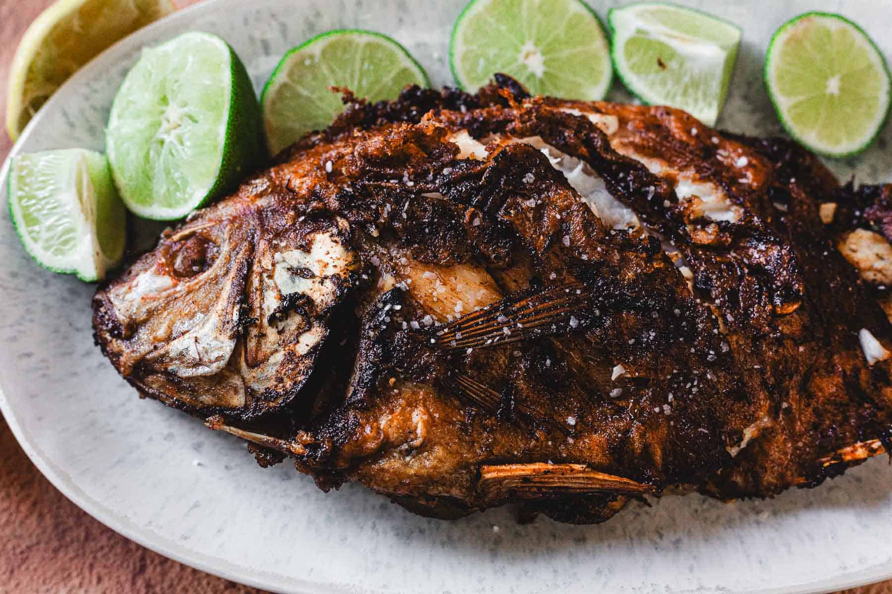 A crispy Whole Fried Mojarra with golden brown skin is served on a white oval plate, accompanied by several halved and sliced limes for a classic Colombian touch.