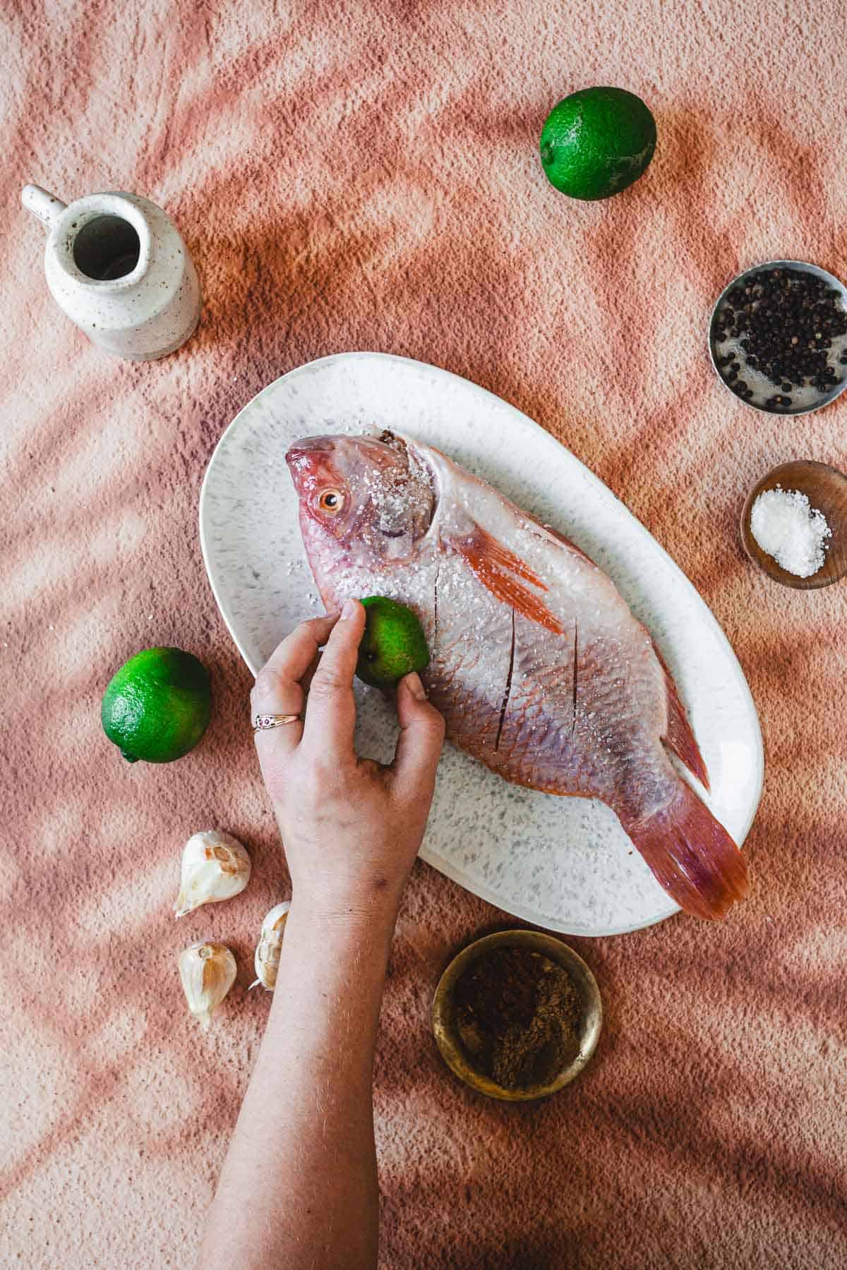 A hand rubs a halved lime onto a raw whole fish—soon to become a delicious Colombian Mojarra Frita—on a white plate, surrounded by whole limes, garlic cloves, salt, pepper, and a small jug on a pinkish textured surface.