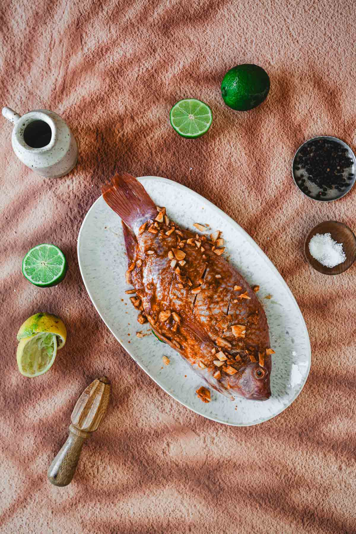 A whole fried Mojarra Frita topped with chopped garlic sits on an oval plate, surrounded by lime halves, a juicer, a small pitcher, a bowl of salt, and pepper on a brown textured surface—classic Colombian flavors.