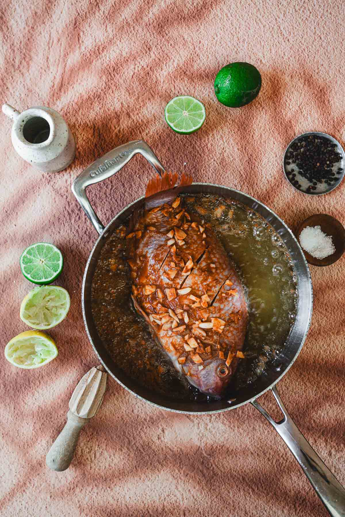 A whole fried mojarra, or mojarra frita, is sizzling in a pan with oil and chopped garlic. Halved limes, a lime squeezer, peppercorns, and salt surround the Colombian dish on a textured surface.