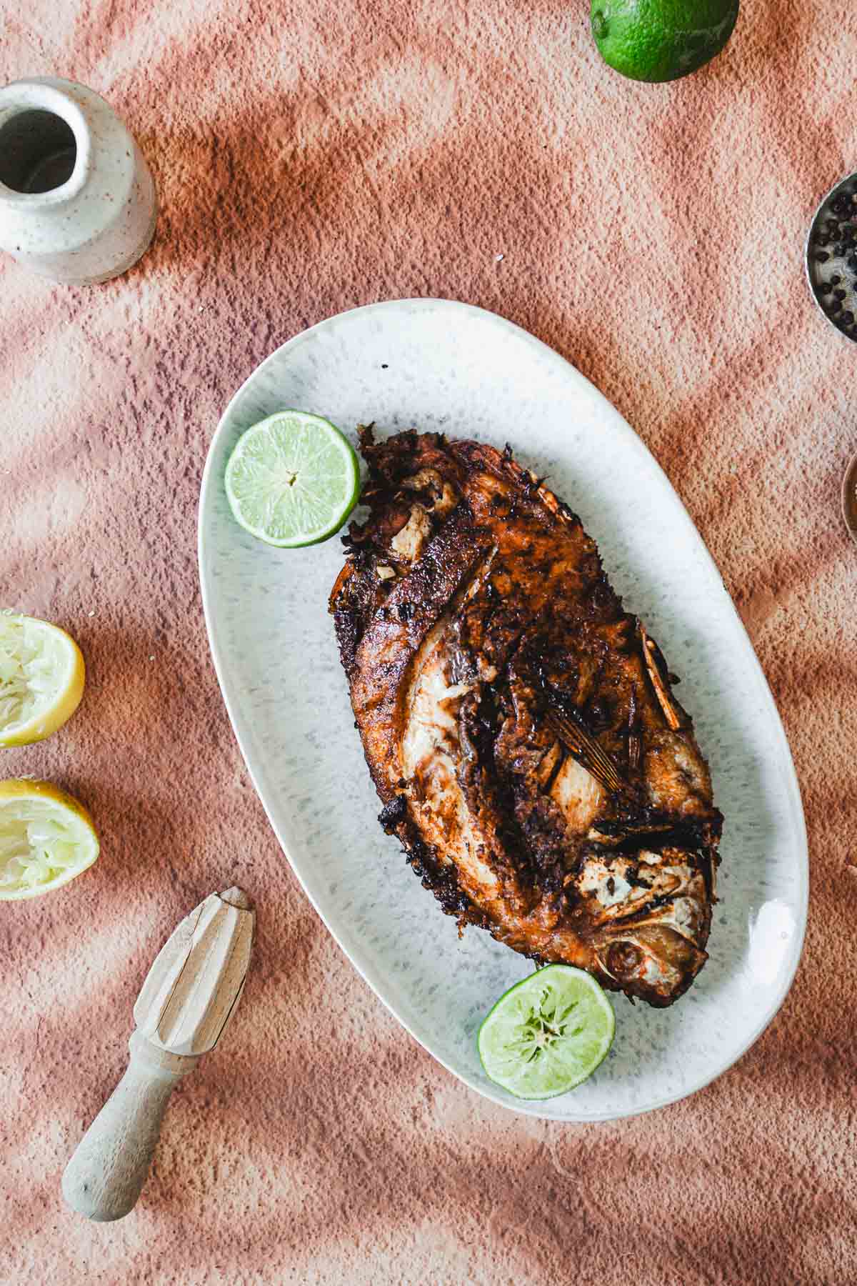 A Colombian Whole Fried Mojarra with a charred exterior is served on an oval plate with two halved limes. The plate rests on a textured peach-colored cloth, surrounded by lemon halves, a citrus juicer, and a small jar.