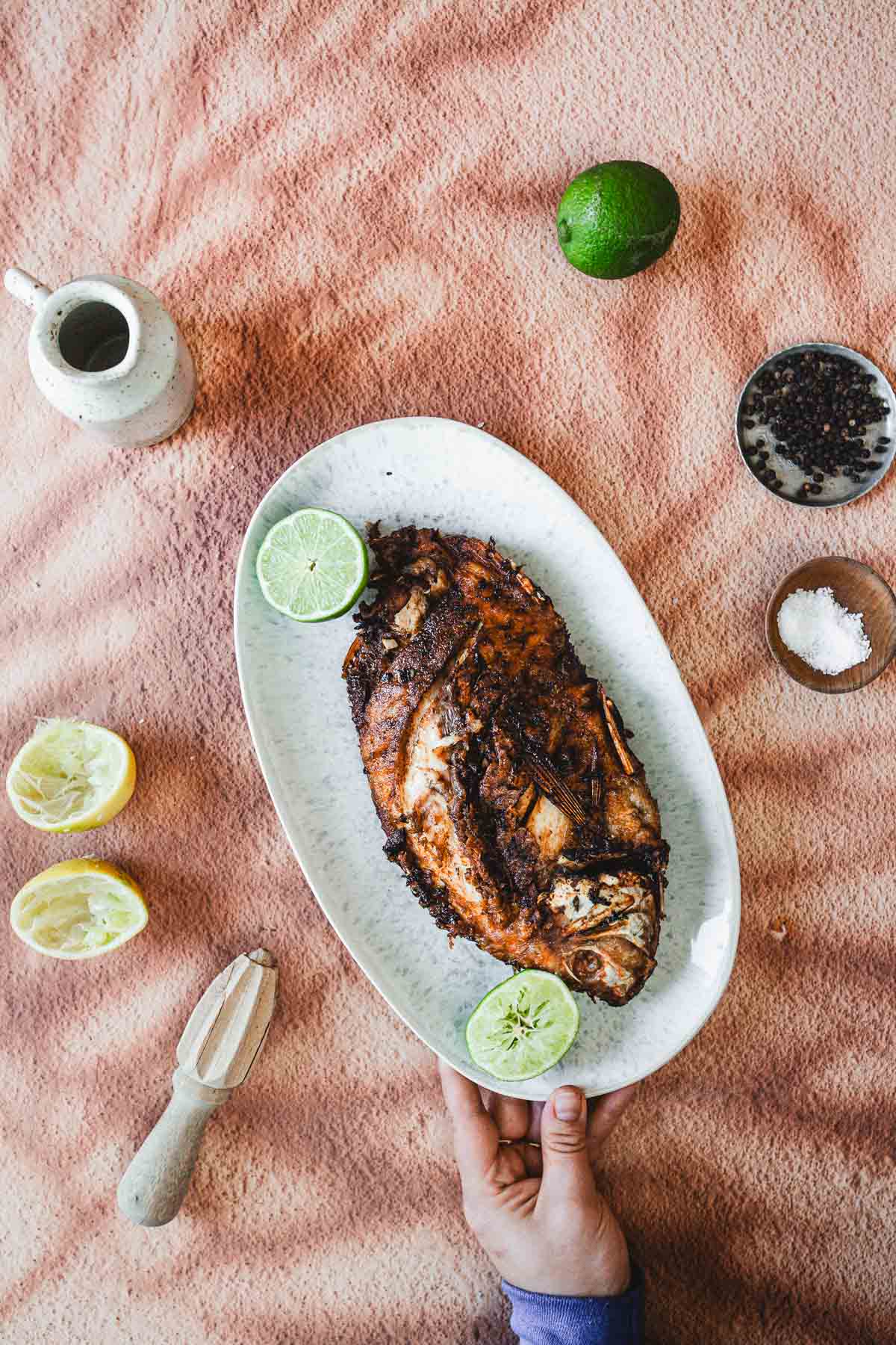 A hand holds an oval plate with a Colombian Whole Fried Mojarra garnished with lime halves. Surrounding the plate are a lime, sliced lemons, a small pitcher, a bowl of peppercorns, and a bowl of salt on peach-colored fabric.