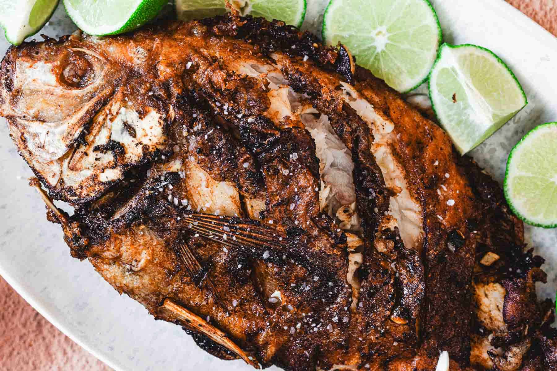 A whole fried mojarra with a crispy, browned exterior is served on a white plate, garnished with coarse salt and several slices of fresh lime—a classic Colombian dish known as Mojarra Frita.