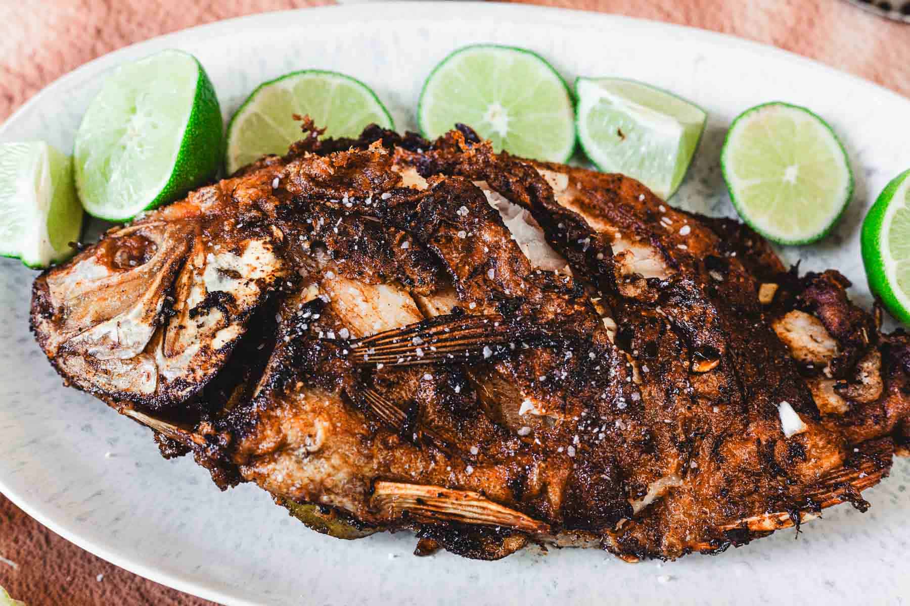 A Whole Fried Mojarra with a crispy, golden-brown exterior is served on an oval plate, garnished with coarse salt and lime wedges—an authentic Colombian frita delight.