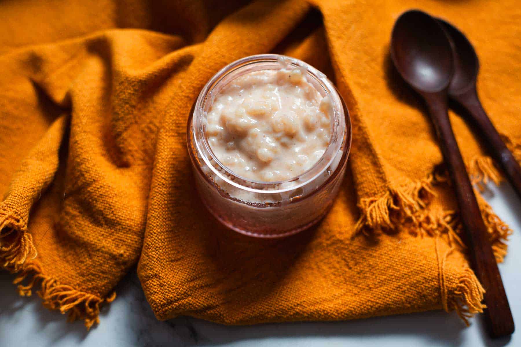 A glass jar filled with creamy Colombian rice pudding sits on an orange textured cloth. Two dark wooden spoons lie to the right of the jar on the cloth. The background is a white surface.