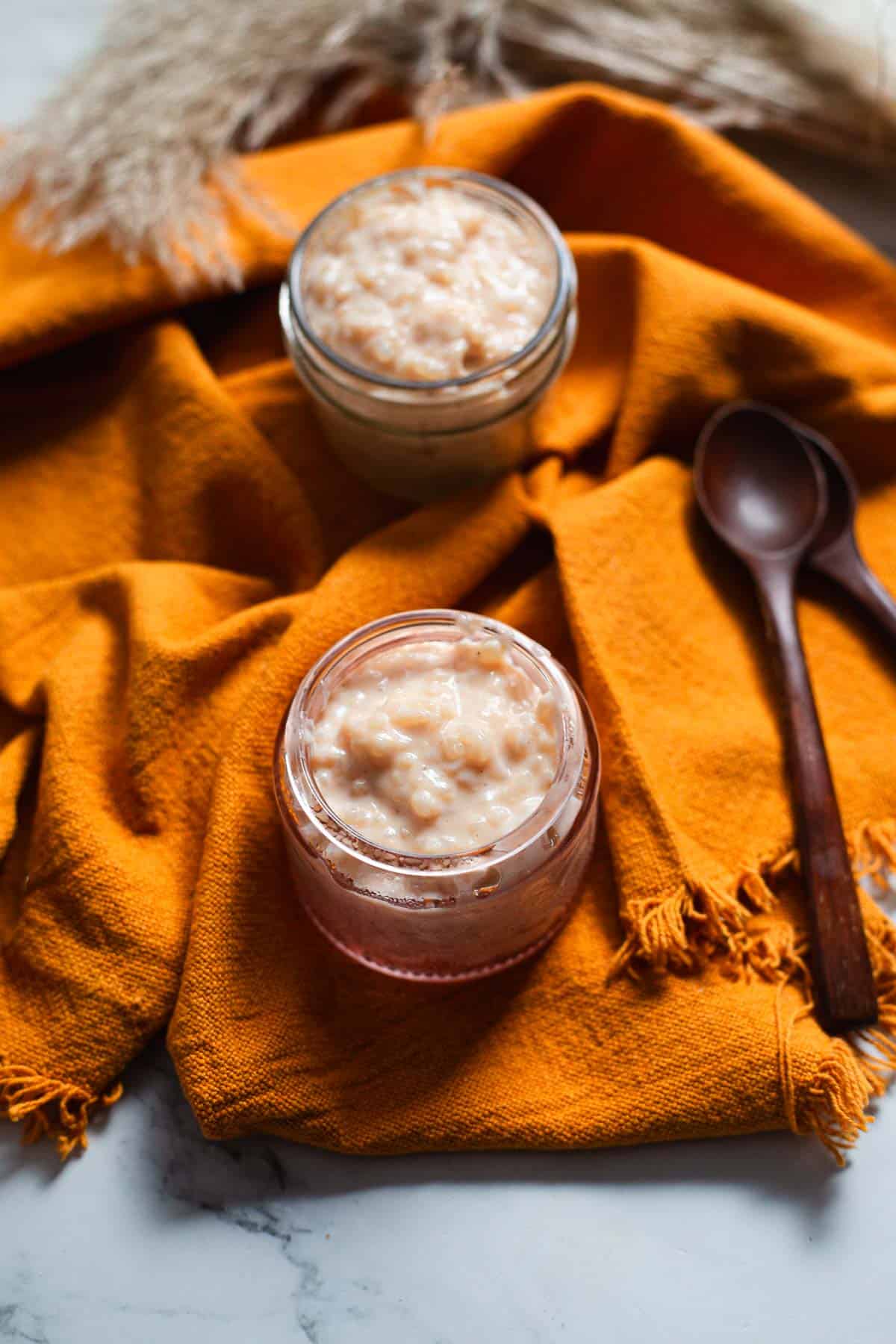 Two small glass jars filled with creamy Colombian rice pudding sit on an orange cloth, with two wooden spoons beside them. Dried grasses are partially visible in the background, highlighting this inviting Colombian dessert.