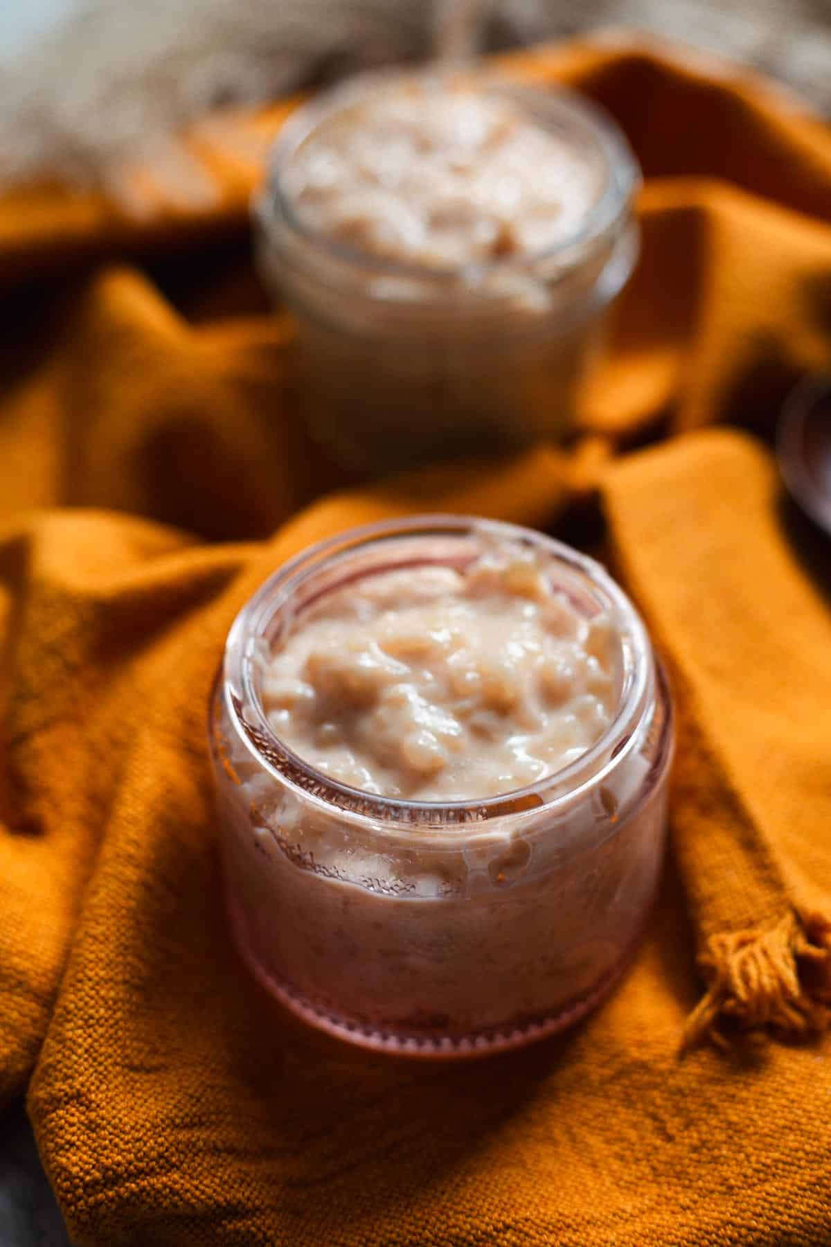 A small glass jar filled with creamy Colombian rice pudding sits on an orange cloth, with another jar of the same dessert blurred in the background.