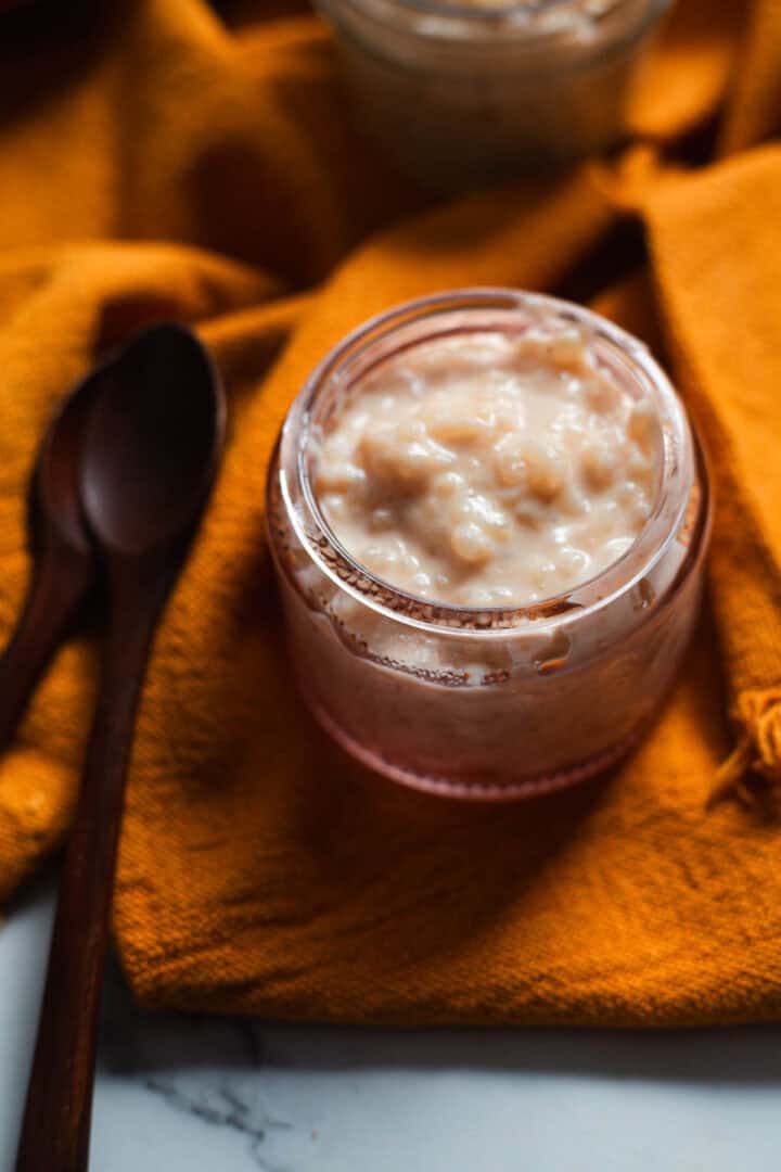A small glass jar filled with creamy Colombian rice pudding sits on an orange cloth, with two dark wooden spoons placed nearby on a marble surface.