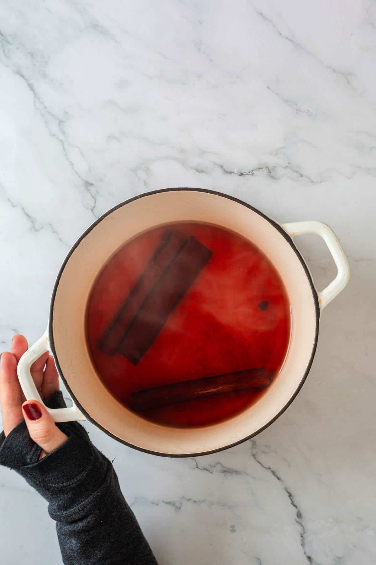 A hand holding the handle of a white pot filled with red liquid and two cinnamon sticks, placed on a white marble surface. Steam is rising from the pot, evoking the warmth and comfort of a classic Colombian dessert like rice pudding.