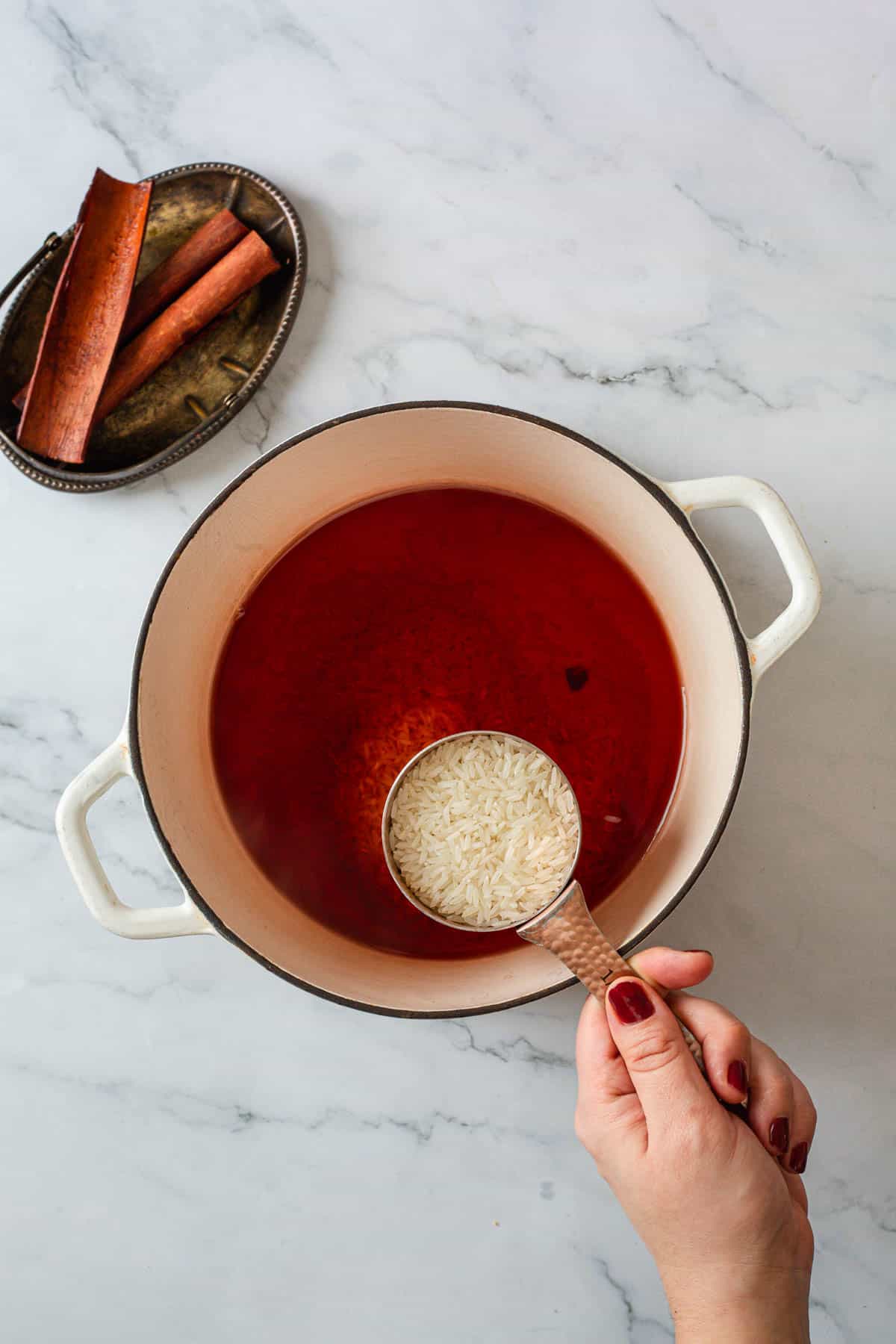 A hand holds a copper measuring cup of uncooked rice over a white pot filled with red liquid, preparing rice pudding. A small tray with cinnamon sticks sits beside the pot on a marble surface, hinting at this Colombian dessert.