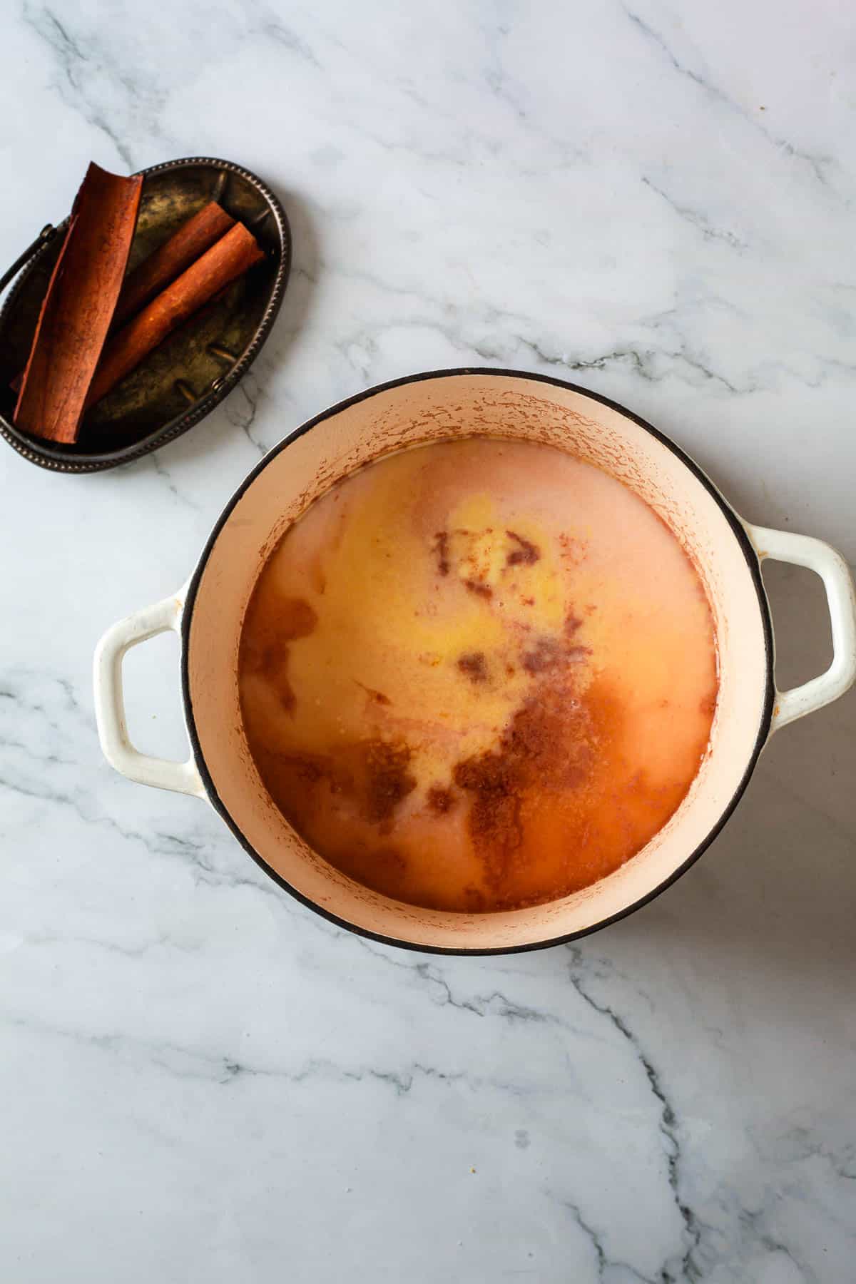 A white pot filled with liquid and visible spices sits on a marble surface, suggesting the beginnings of Colombian rice pudding. Next to it is a small metal tray holding several cinnamon sticks.