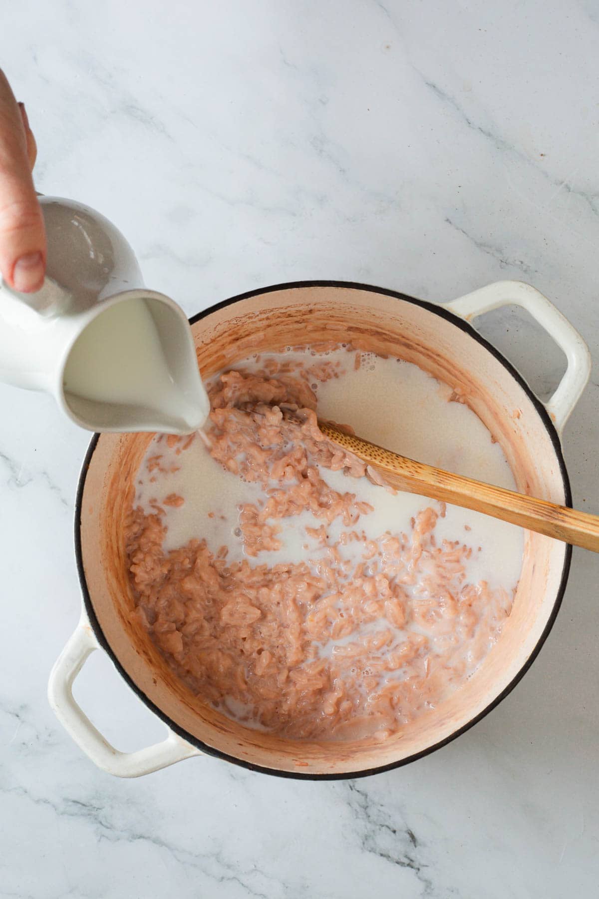 A hand pours milk from a small white pitcher into a pot of partially cooked Colombian Rice Pudding while a wooden spoon rests in the mixture. The pot sits on a marble countertop.