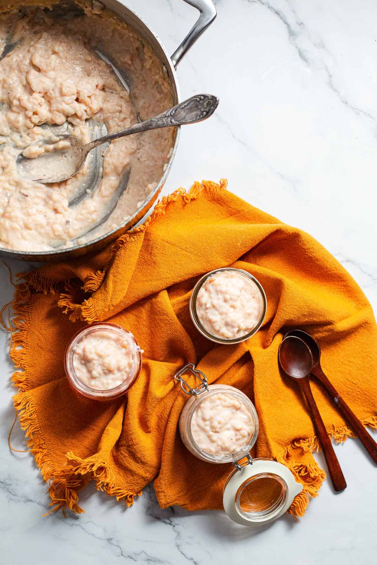 A saucepan with Colombian Rice Pudding and a spoon sits on a marble surface next to three jars filled with this classic Colombian dessert, all resting on an orange cloth alongside two wooden spoons.