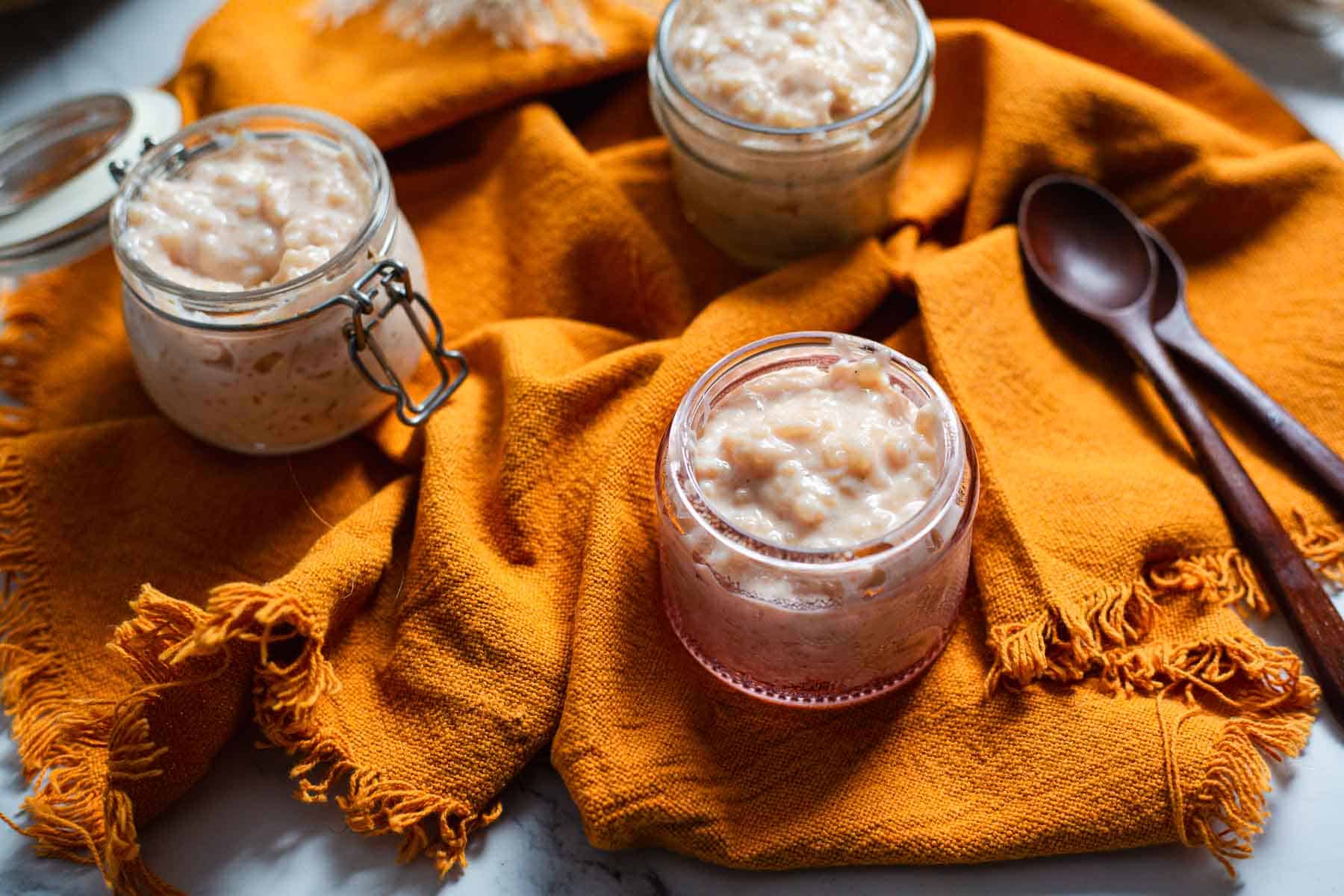 Three small glass jars filled with Colombian rice pudding sit on an orange textured cloth. Two brown spoons are placed nearby, and the jars are arranged in a triangular pattern on a marble surface.