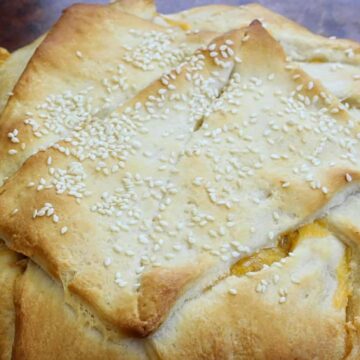 A round baked pastry with a golden-brown crust, topped with sesame seeds. The pastry appears folded with visible seams and some yellow filling peeking out from the edges.