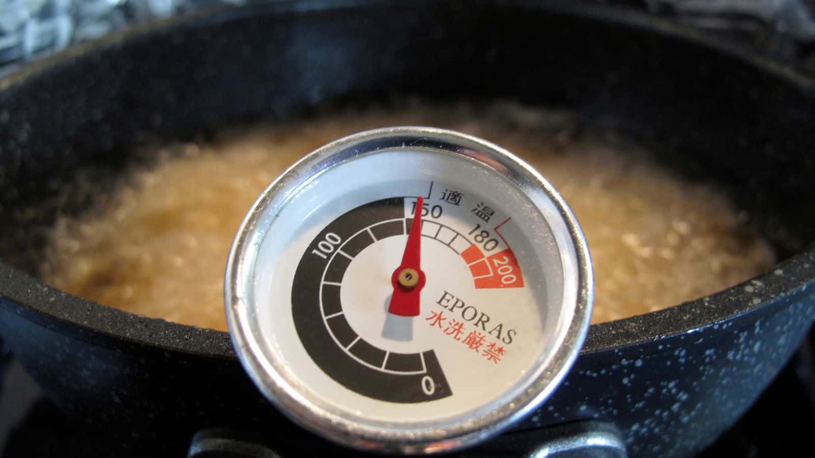 A close-up of a thermometer in a black pot with liquid boiling inside. The thermometer needle points to around 170°C, and the dial has markings in both numbers and Japanese characters.
