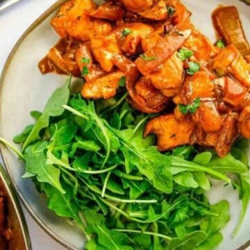 A plate of cooked chicken pieces in a reddish sauce with herbs, served next to a generous portion of fresh arugula leaves. A fork and a pan with more chicken are partially visible nearby.