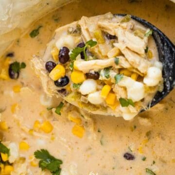 A close-up of a creamy soup with shredded chicken, black beans, corn, hominy, and chopped herbs, being ladled from a pot lined with parchment paper.