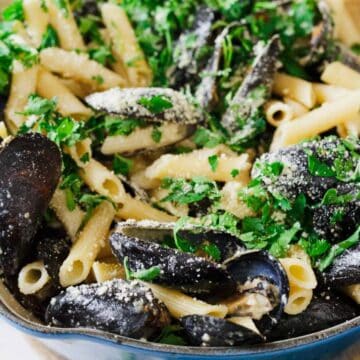 Pasta with cooked mussels, garnished with chopped parsley and grated cheese, is served in a large blue pot. Some fresh parsley is visible in the background.