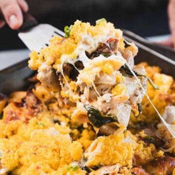 A person lifts a portion of cheesy baked pasta with vegetables and a golden breadcrumb topping from a rectangular baking dish using a spatula.