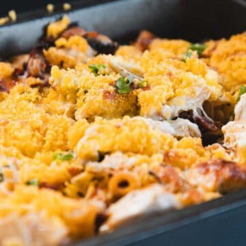 A close-up of a baked casserole in a rectangular metal pan, topped with a golden breadcrumb or cheese crust, with visible pasta and pieces of chicken or vegetables scattered throughout.