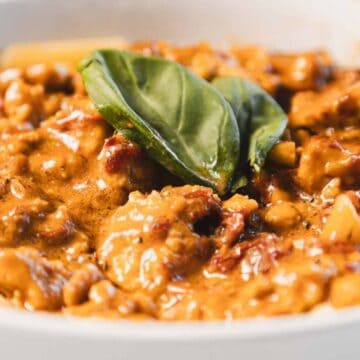 A close-up of rigatoni pasta topped with a creamy tomato-based sauce containing chunks of meat and garnished with a fresh basil leaf, served in a white bowl.