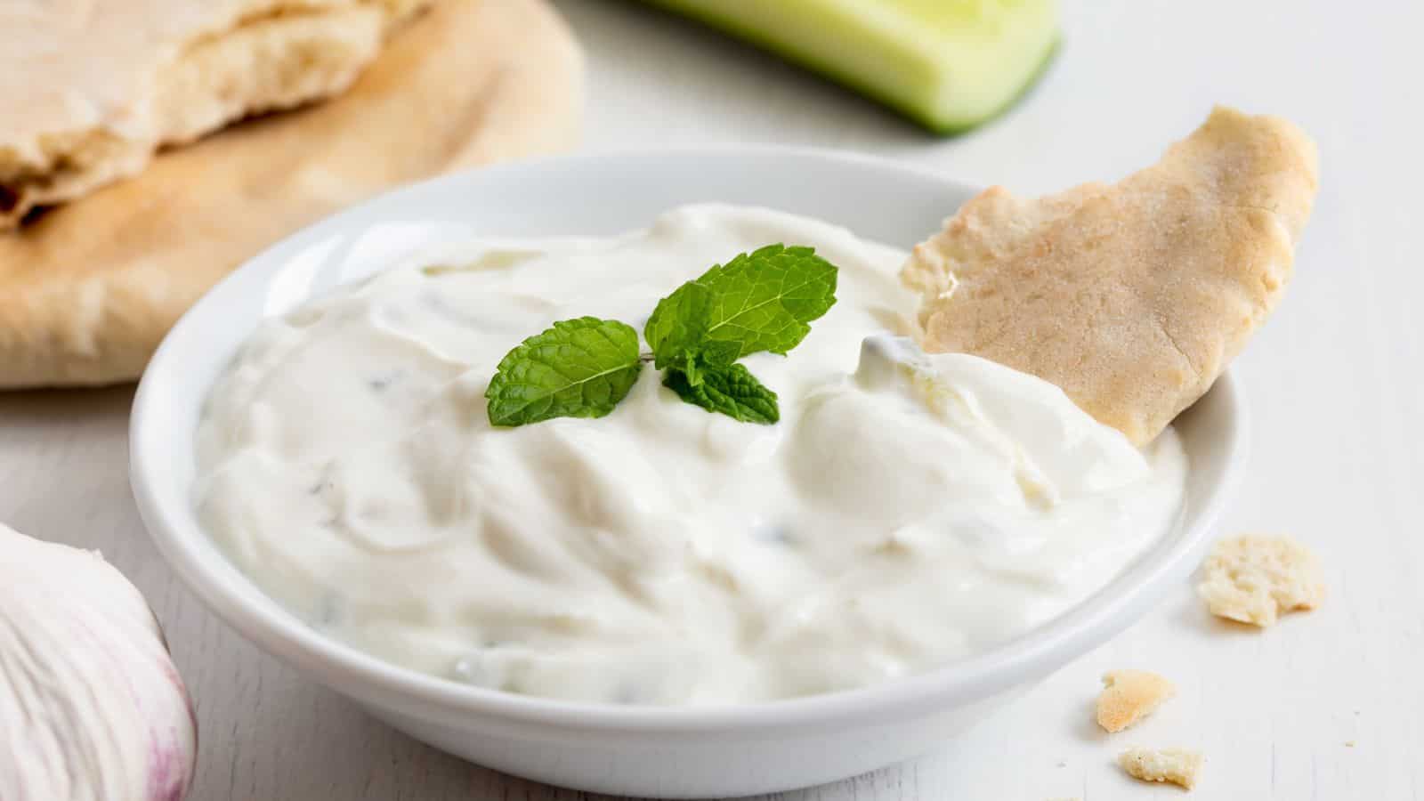 A white bowl of creamy tzatziki dip garnished with two mint leaves, with a piece of pita bread partially dipped in it. Some pita bread and a cucumber slice are in the background on a white surface.