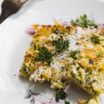 A slice of vegetable frittata topped with grated cheese and fresh parsley sits on a floral-patterned plate, with a fork beside it and some parsley garnish on the plate.