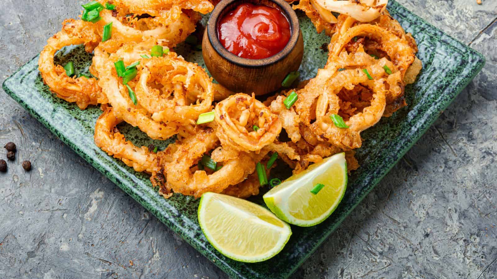 A rectangular green plate with fried calamari rings topped with chopped green onions, two lime wedges, and a small bowl of red dipping sauce placed in the center. The plate is on a gray textured surface.