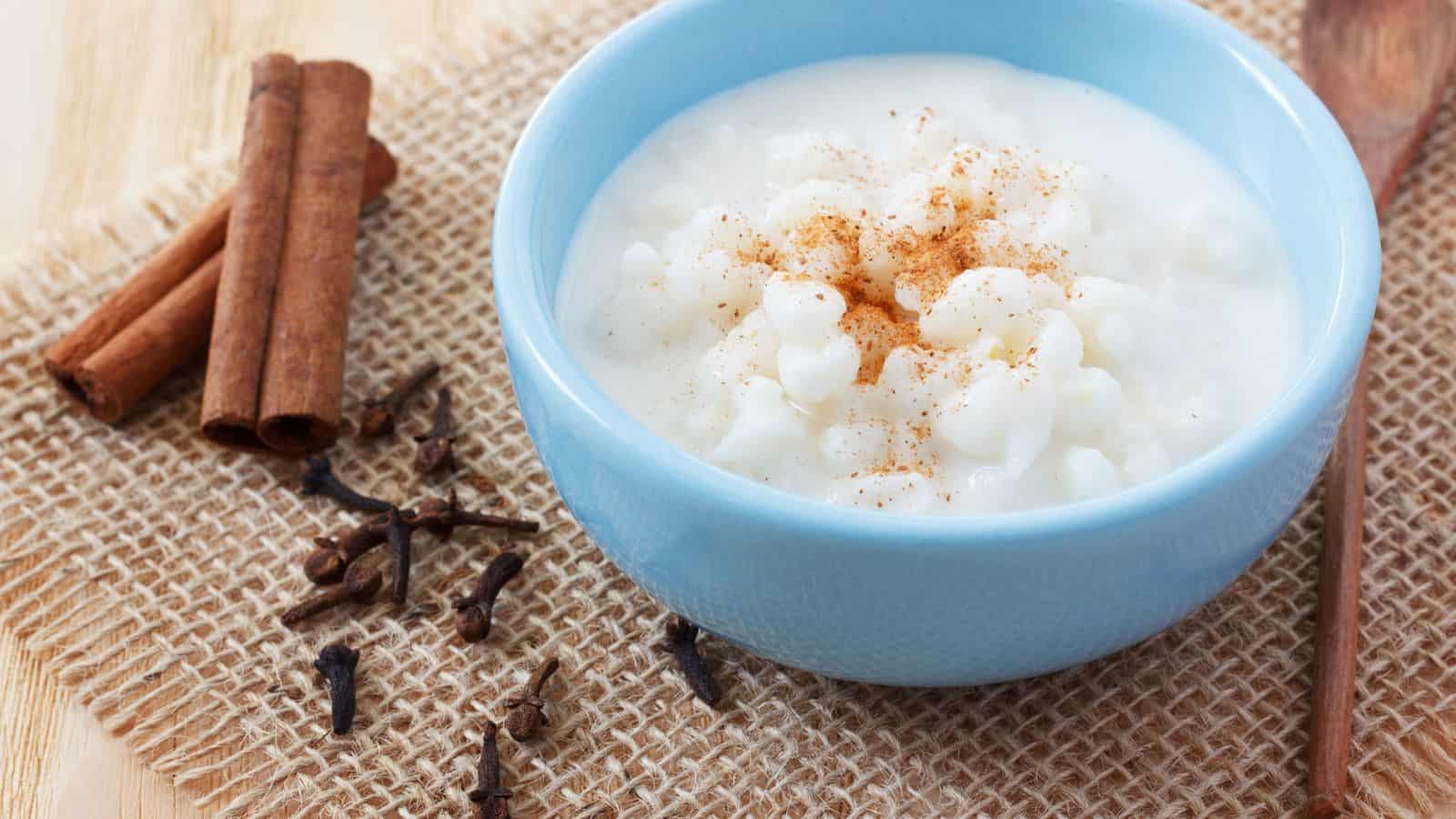 A blue bowl filled with rice pudding topped with ground cinnamon sits on a piece of burlap. Beside the bowl are whole cinnamon sticks, cloves, and a wooden spoon.