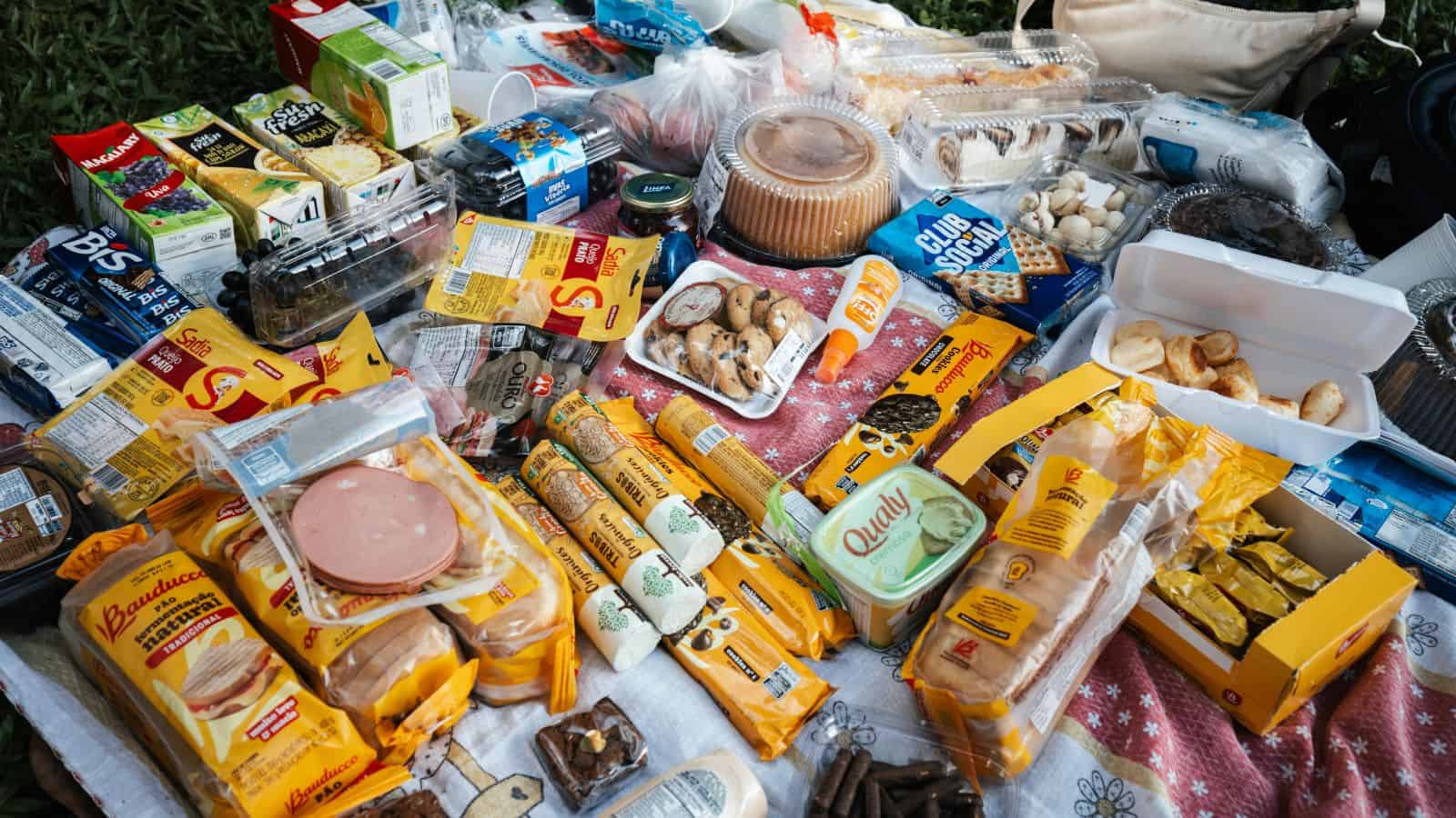 A picnic spread on a patterned cloth featuring packaged bread, pastries, cakes, cold cuts, juice cartons, cookies, cheese, canned goods, plastic containers with food, and other assorted snacks and drinks.