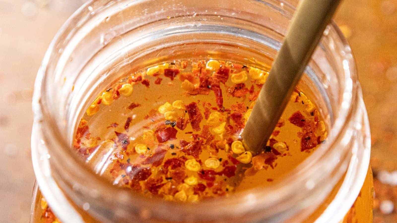 Close-up of a glass jar filled with liquid containing chili flakes. A spoon is dipped into the mixture, and the vibrant red and orange colors of the chili stand out against the clear liquid.