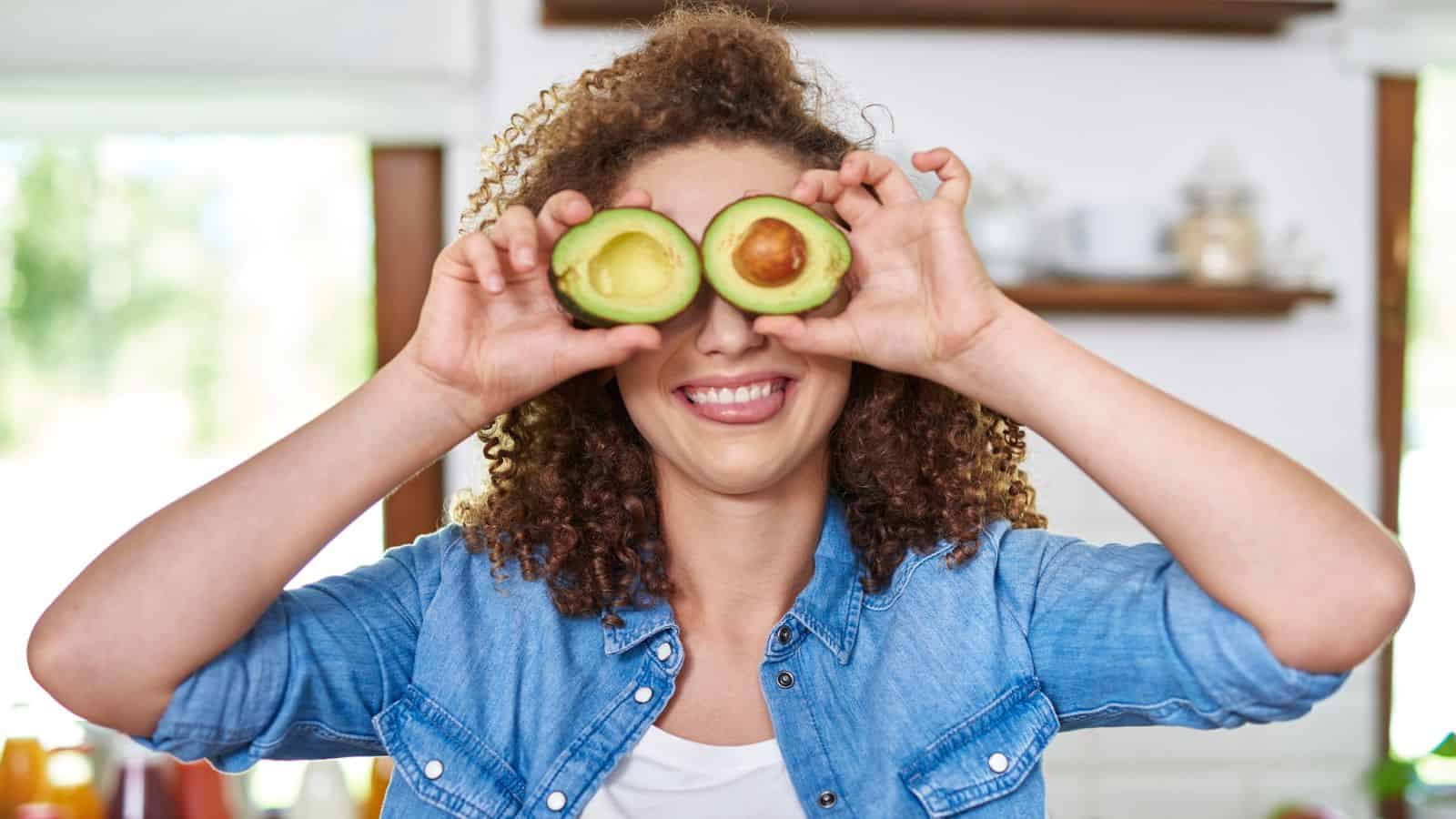 A woman with curly hair holds two avocado halves in front of her eyes while smiling, standing in a bright kitchen.