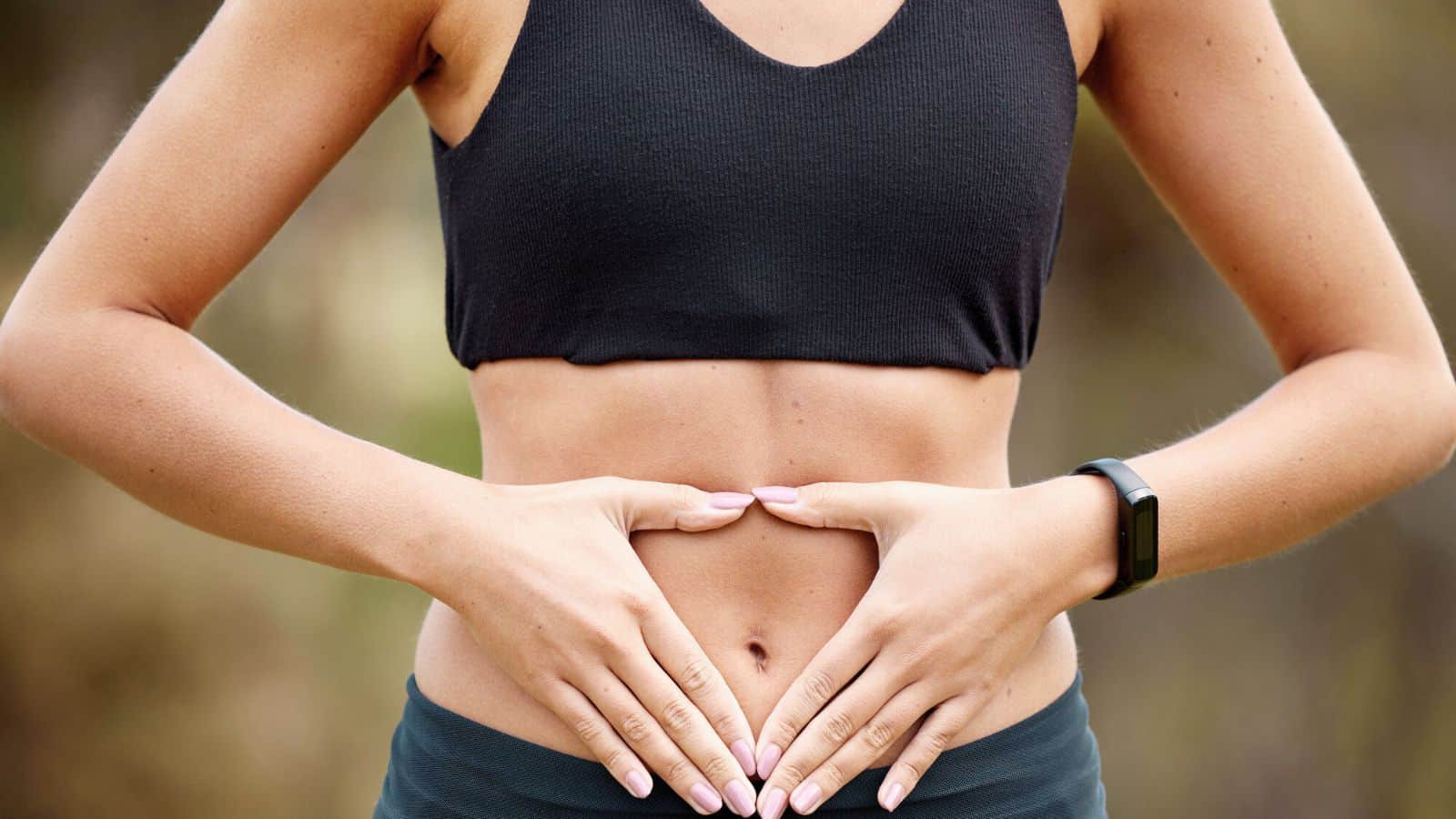 A person wearing a black sports bra and fitness tracker holds their hands in a heart shape over their bare stomach, outdoors. The photo is cropped to show the torso only.