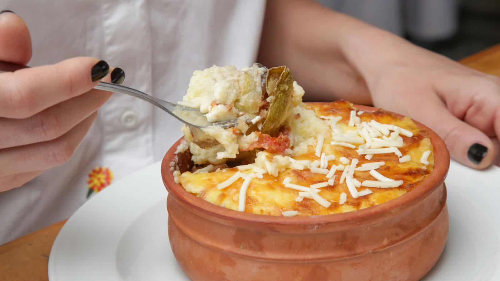 A person holds a spoonful of casserole topped with shredded cheese over a brown ceramic dish containing the baked casserole, which sits on a white plate.