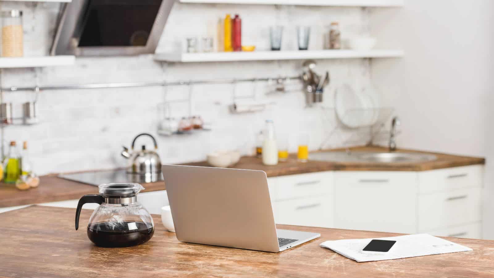 A modern kitchen with a wooden countertop featuring a laptop, a glass coffee pot, a smartphone, and papers. The background shows a sink, kettle, utensils, and shelves with various items.