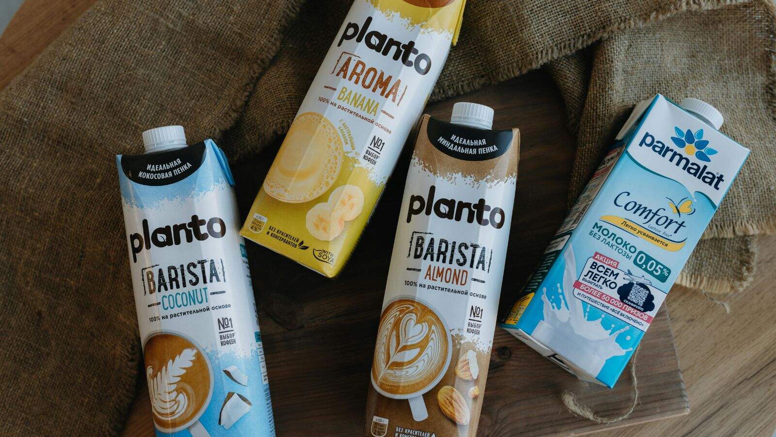 Four cartons of drinks are arranged on a wooden surface. Three are plant-based "Planto" beverages in coconut, almond, and banana flavors, and one is a blue and white "Parmalat Comfort" milk carton with Russian text.