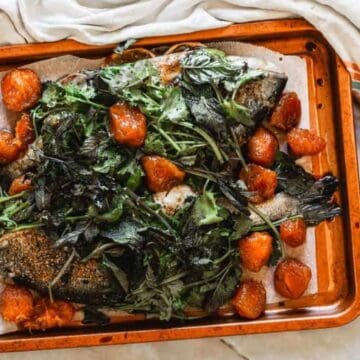 A roasted whole fish topped with leafy greens and roasted tomatoes on a baking tray lined with parchment paper, next to a bowl of peppercorns and a striped cloth.