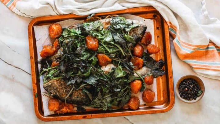A roasted whole fish topped with leafy greens and roasted tomatoes on a baking tray lined with parchment paper, next to a bowl of peppercorns and a striped cloth.